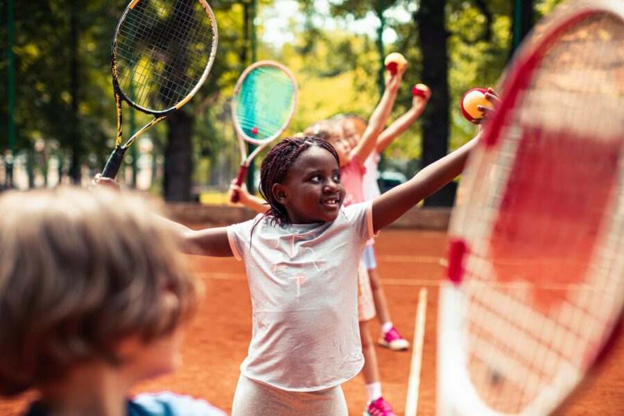 Image of children playing tennis outdoors in the sunshine. The children are standing in a row holding up their tennis balls and rackets.