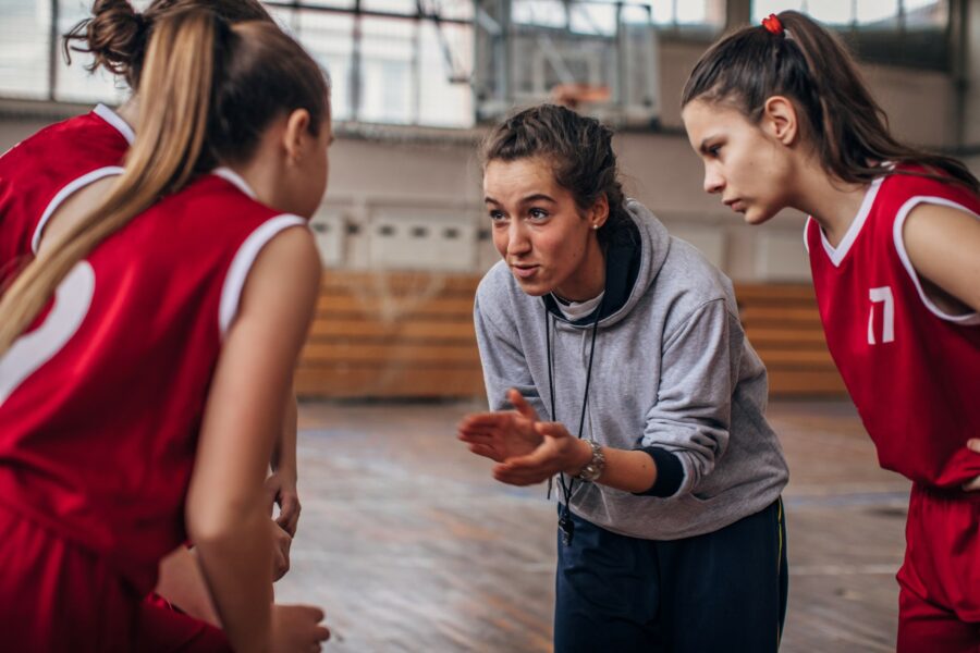 A young female coach, she is wearing a grey hoodie navy tracksuit trousers. The coach appears to be giving instructions to her netball players, she is leaning into the huddle and clapping her hands in an encouraging gesture. There are three players leaning in towards the coach, listening closely to what she is telling them. The players appear to be teenaged females, and they are all dressed in the same kit, indicating that they are on the same team. Their kit consists of a sleeveless red top with white trim and red shorts. They are standing in a school sports hall setting and there is a basketball hoop in the background.