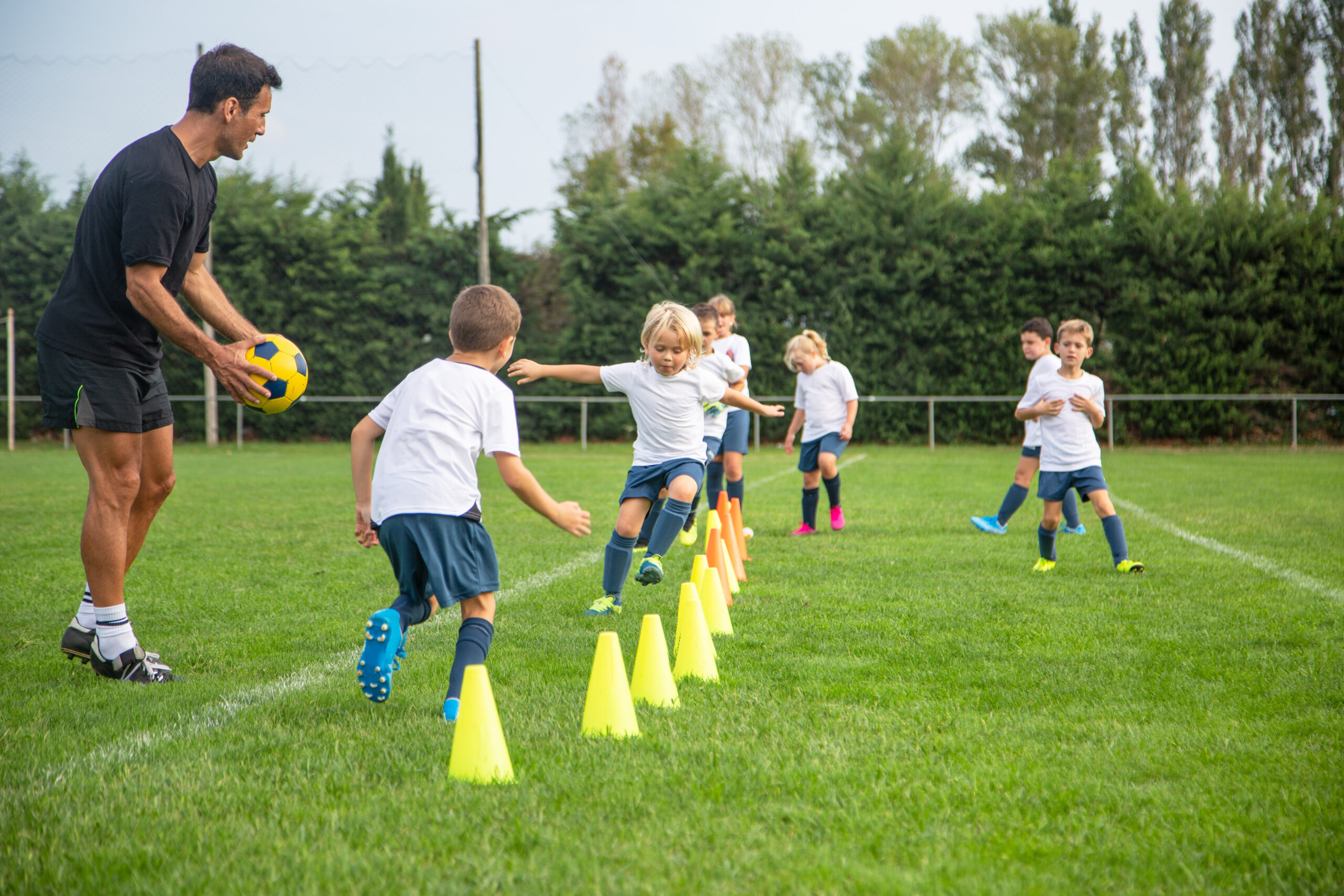 Image showing a group of young footballers practicing running drills practice, outdoors on a field. The adult football coach appears to be on the left hand side of the image and is holding a football. The children are all dressed in white and blue t-shirts and shorts.